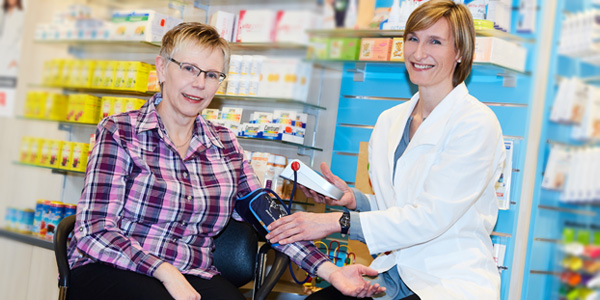 Blutdruckmessung in einer Apotheke: Eine sitzende Person erhält mit einer Blutdruckmanschette am Arm eine Messung durch eine Person in weißer Arbeitskleidung. Im Hintergrund sind Apothekenregale mit Produkten sichtbar.